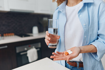 Woman Holding Glass of Water and Pill in Hand Standing in Front of Kitchen Counter