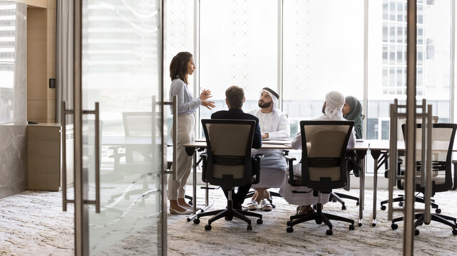 Arabian and European businesspeople discussing collaboration in modern office business meeting room, sitting at large table, talking to standing female group leader. Full length banner shot