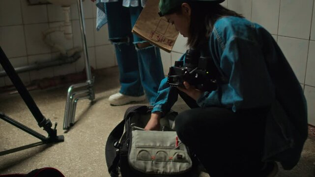 Side tilt shot of young Caucasian lady taking video camera out of bag while preparing movie set before shooting movie in washroom, working on final project with diverse group mates