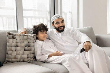 Cheerful Muslim father and positive son boy watching TV show, movie, series, video together, setting channel with remote control, enjoying leisure on home couch with online media service