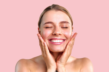 Satisfied happy smiling young woman touching her glowing skin and smiling on soft pink background. The concept of advertising, skin care, cosmetics.