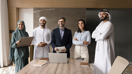 Multiethnic of happy Arabic and European business leaders, partners, entrepreneurs standing together at office large table, smiling, looking away, posing for group portrait after meeting