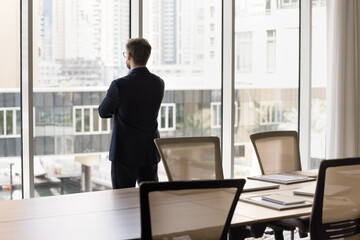 Pensive confident businessman looking out of window in deep thoughts, standing at glass with arms...