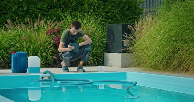 Pool technician checking data on a tablet beside a swimming pool with chemical containers and lush greenery.