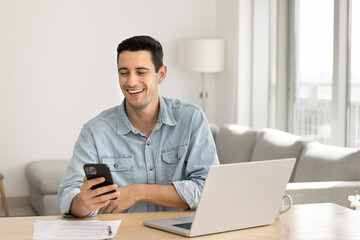 Happy successful Latin freelance businessman using digital gadgets for professional communication from home, holding mobile phone, chatting, smiling at screen, sitting at laptop in apartment