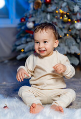 Happy toddler sitting on plush rug near decorated Christmas tree. A smiling toddler plays on a soft rug, surrounded by a beautifully adorned Christmas tree