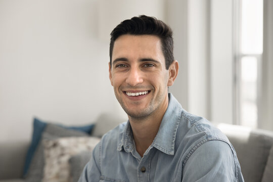 Happy handsome young Hispanic man in casual denim shirt looking at camera with toothy smile, opening white teeth, sitting on couch at home, posing for portrait, feeling cheerful, positive