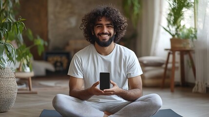 Cheerful sporty millennial indian guy with beard and curly hair sitting on yoga mat showing modern smartphone with empty screen recommending nice fitness mobile app bedroom interior mo : Generative AI