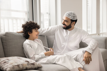 Happy Arabian son kid and loving dad in white Muslim dresses talking and laughing on home couch, having fun, enjoying family leisure, good close relationship, conversation