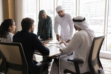 Business team of young serious Arabic managers discussing teamwork, project strategy with Arabian boss and European partners at meeting, standing at table, speaking to sitting colleagues