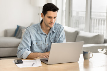 Focused young Latin freelance business owner man working at home computer, typing on laptop, sitting at table, with cozy living room in background, using desktop for work on project