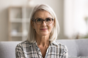 Head shot close up portrait attractive mature woman in eyewear sit on sofa, look at camera spend time at home. Older generation person advertises eyesight check-up, lenses store, professional services