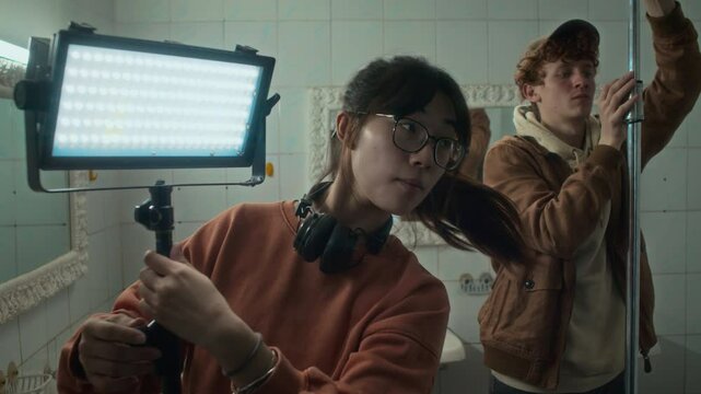 Chest up shot of Asian lady in glasses and longsleeve adjusting mini floodlight while preparing film set