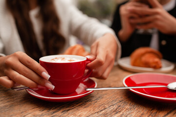  girls are sitting in a French cafe, eating a croissant and drinking coffee with whipped cream. Teenagers are dressed in business style, black and white jacket close-up of the hands holds red cup