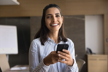 Cheerful beautiful Hispanic businesswoman holding smartphone, posing with gadget in office boardroom in boardroom, looking at camera, smiling, typing on mobile phone. Office portrait