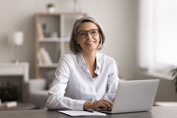 Happy middle-aged woman with short gray hair and glasses, smile, look at camera, seated in cozy...