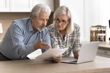 Serious worried older couple read legal documents letter, checking domestic bills stand at kitchen...
