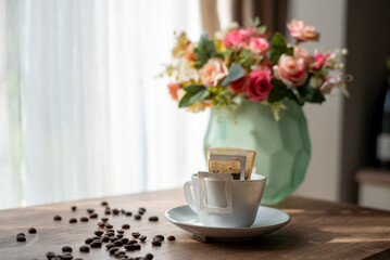 Morning coffee filled with drip coffee filters on wooden table. Hot black coffee cup, with bouquet of flowers background surrounded by scattered coffee beans on desk.