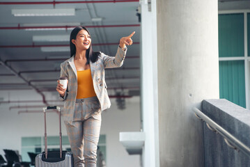 Confident businesswoman in stylish plaid suit point and holding coffee cup, ready to start journey. Woman stands in airport terminal, with carry-on suitcase beside her. Business Travel concept.