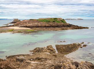 Coastal landscape of Saint Malo in Brittany with jagged rocks, sandy beach, cloudy sky, and small...