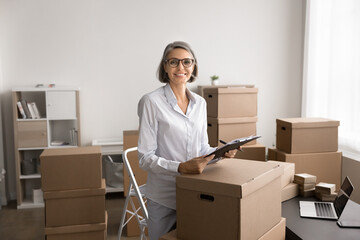 Mature woman standing in office space surrounded by cardboard boxes and prepared packages for...