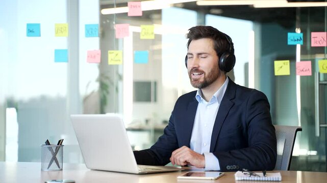 Happy businessman wearing wireless headphones listening to music during a break in work on a laptop sitting at workplace in a business office. Joyful male closes his eyes and enjoys track singing song