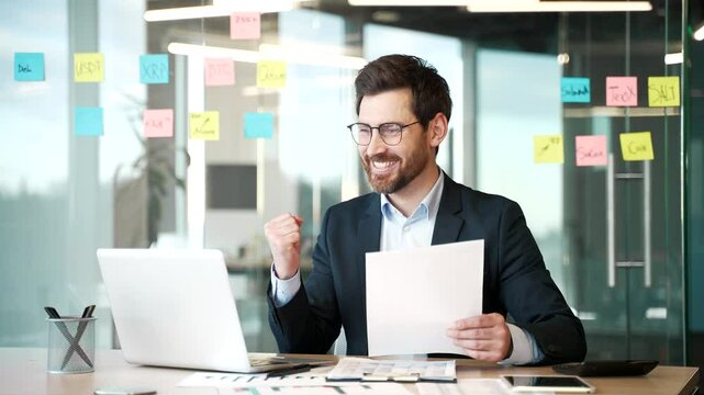 Happy financier satisfied with financial results sitting at workplace in business office. Smiling satisfied businessman investor looks at documents and computer and celebrates successful investment