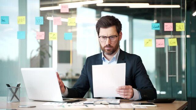 Confused businessman in a formal suit having difficulty with paper work checking documents on the computer sitting at workplace in business office. Frustrated financier unhappy with financial results