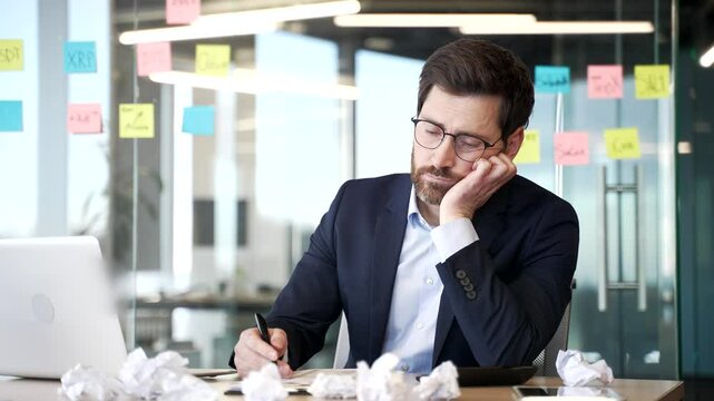 Overworked bearded businessman is bored sitting at workplace at desk in business office. Exhausted financier is tired of doing paper work. Man the owner has depression, despair. Crisis and failure