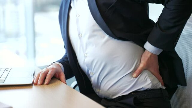 Close up. Male hand holding massaging lower back. Man in a formal suit suffering from back pain sitting at the desk at workplace in business office. Businessman massages painful muscles, sick spine
