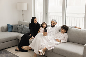 Happy Arabic parents and two little children using smartphone together, enjoying online communication, shopping, booking on Internet, making order. Family resting with cellphone on home couch