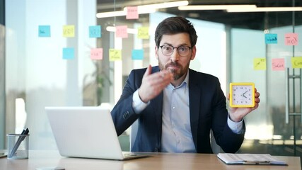Worried businessman in suit holding clock with urgent expression in and shows looking at camera while sitting at desk at workplace in modern business office. Concept of time management and deadlines