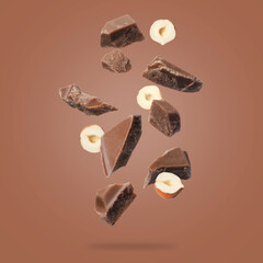 Pieces of tasty chocolate and hazelnuts falling on pale brown background