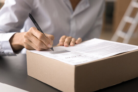 Cropped close up shot unknown businesswoman labeling or signing package box seated at her desk. Logistics management, order fulfillment, or inventory tasks in a small business or home office setting