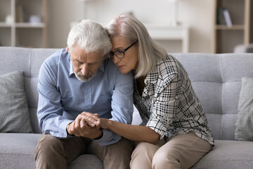 Loving older wife comforting her upset husband, sitting on sofa hugging his, provide psychological...