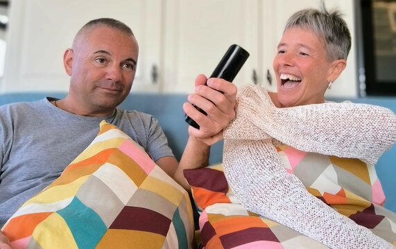 Happy couple sitting on sofa with remote control in hand and watching tv together.Smiling boyfriend and girlfriend joking while watching tv,sitting on sofa.Couple having fun sitting on sofa watching