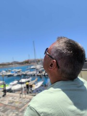 Young adult man sitting at the port,seen from behind looking up. Man from behind looking at the sky,sitting at the port.Tourist sitting at the pier watching the seagulls fly.White man looking up 