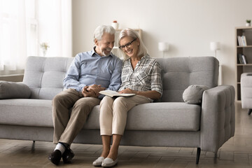 Smiling mature spouses spend peaceful pastime reading a book, enjoy plot of fascinating literature,...