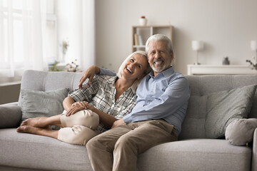 Cheerful mature relaxed spouses posing for camera seated on comfortable couch in modern living room. Baby boomer generation couple photoshoot, harmonic relationship, happy lifelong marriage, bonding