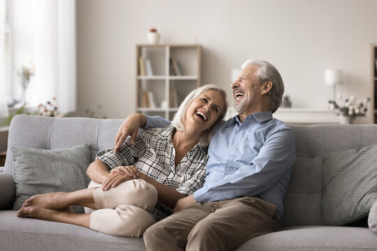Joyful mature wife and husband resting together on couch, hugging, talking and laughing enjoy time together, joking, having fun, relish their harmonic relations, understanding and joint sense humour - Powered by Adobe
