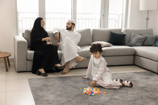Middle Eastern mother and father in Arab clothes talking on cozy sofa, discussing upbringing of little son. Boy sitting on floor, playing building blocks in comfortable home interior. Full body shot - Powered by Adobe