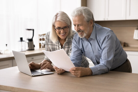 Happy elderly couple hold documents papers, read bank notice with positive news, make payment on internet banking services. Enough of money to pay bills, repayment, pension raise and good fixed income