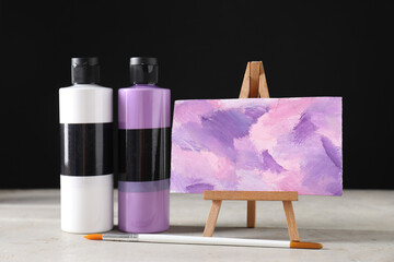 Acrylic paints of different colors in bottles, brush and small easel with abstract picture on light...