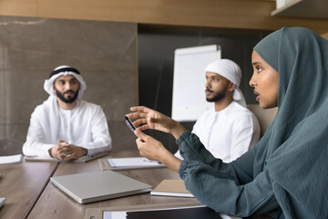 Serious beautiful Arab businesswoman wearing hijab, talking to male colleagues in kanduras at...