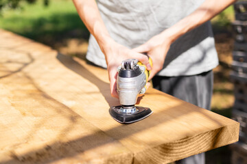 Carpenter Is Using An Electric Sander On A Wooden Plank, Smoothing The Surface For A Woodworking Project Outdoors. Woman Does Physical Work. Concept Of Gender Equality At Work.