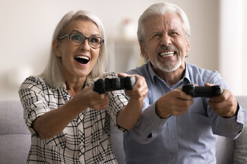 Amazed happy aged married couple having fun at home, holding video game controller, fighting in virtual battle, laughing, playing videogames with excitement and passion. Indoor activity, entertainment