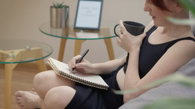 A woman is calmly writing in a notebook, sipping coffee in a comfortable living room. The scene exudes coziness and relaxation, depicting a casual indoor setting for journaling and selfcare