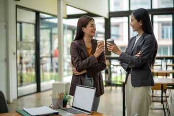 Two women are sitting at a desk with a laptop and a cup of coffee