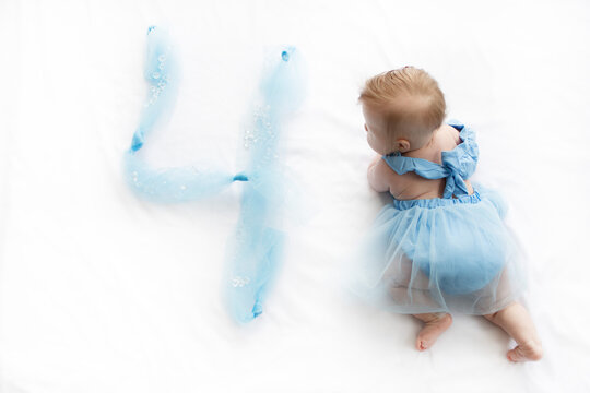 Cute baby girl in blue dress stands near large number four painted on white surface with blue fabric celebrating birthday milestone, symbolizing growth, childhood photography, special memory. - Powered by Adobe
