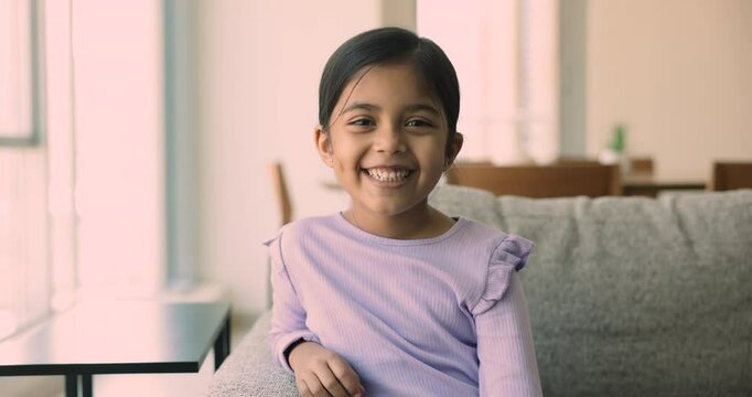 Serious beautiful little Indian girl posing for portrait on home sofa, getting positive, happy, looking at camera with toothy smile, laughing out loud, enjoying childhood